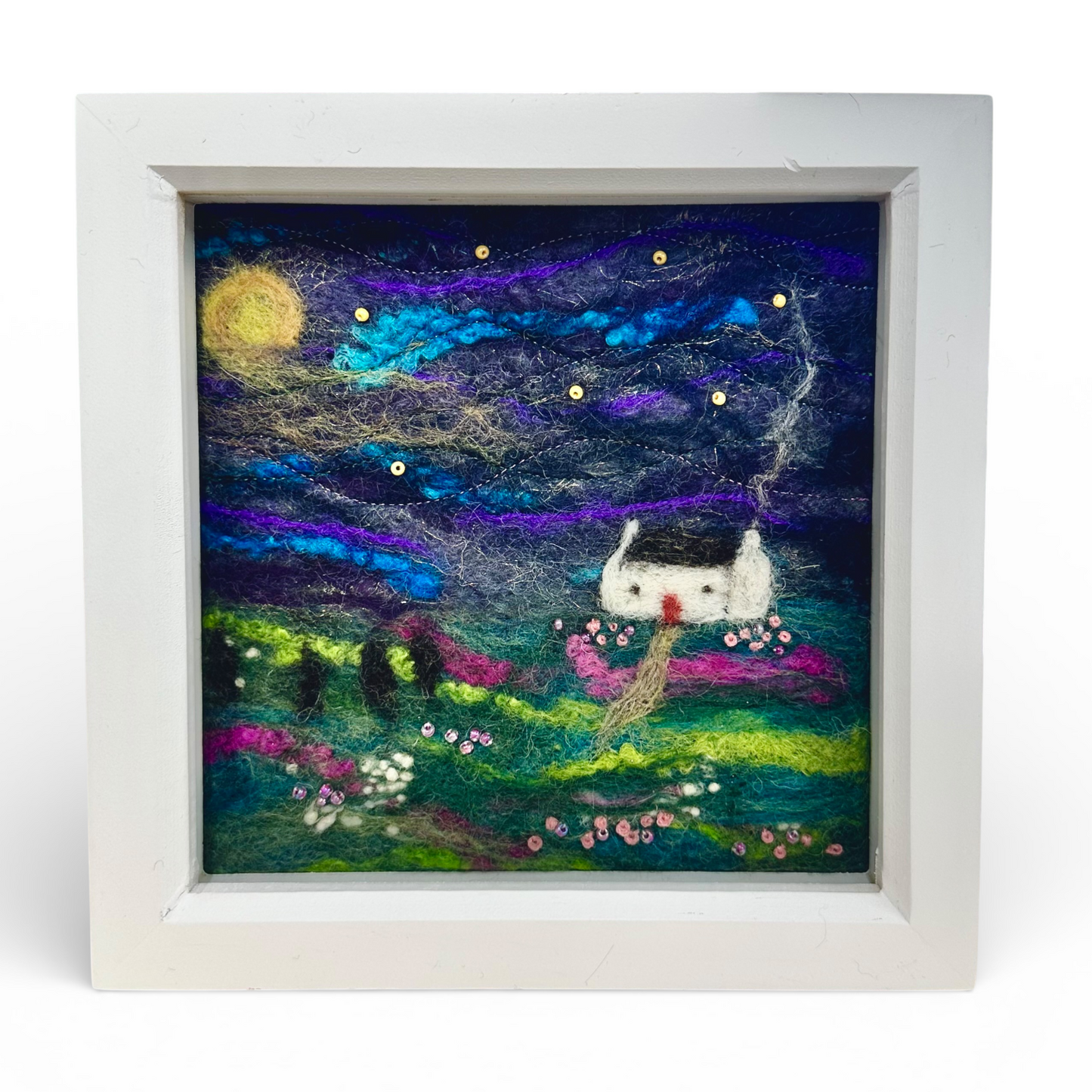 Hand felted wool artwork of a croft house under the northern lights in a white square frame.