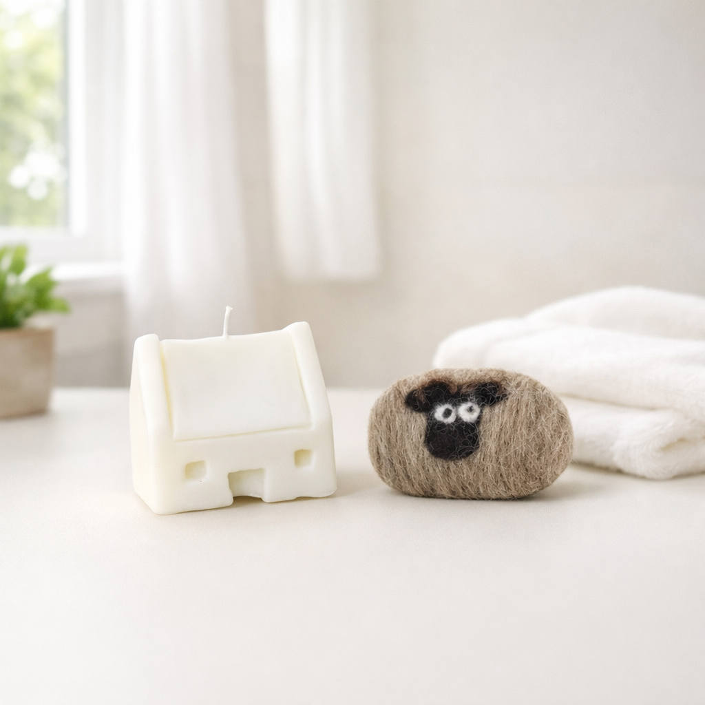 A house-shaped white candle sits next to a brown felted soap with a sheep face design.