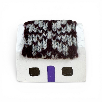 Handmade white croft house with a purple door and a grey and black knitted wool roof.