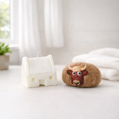 A house-shaped white candle and a brown felted soap with a highland cow face on a white surface.