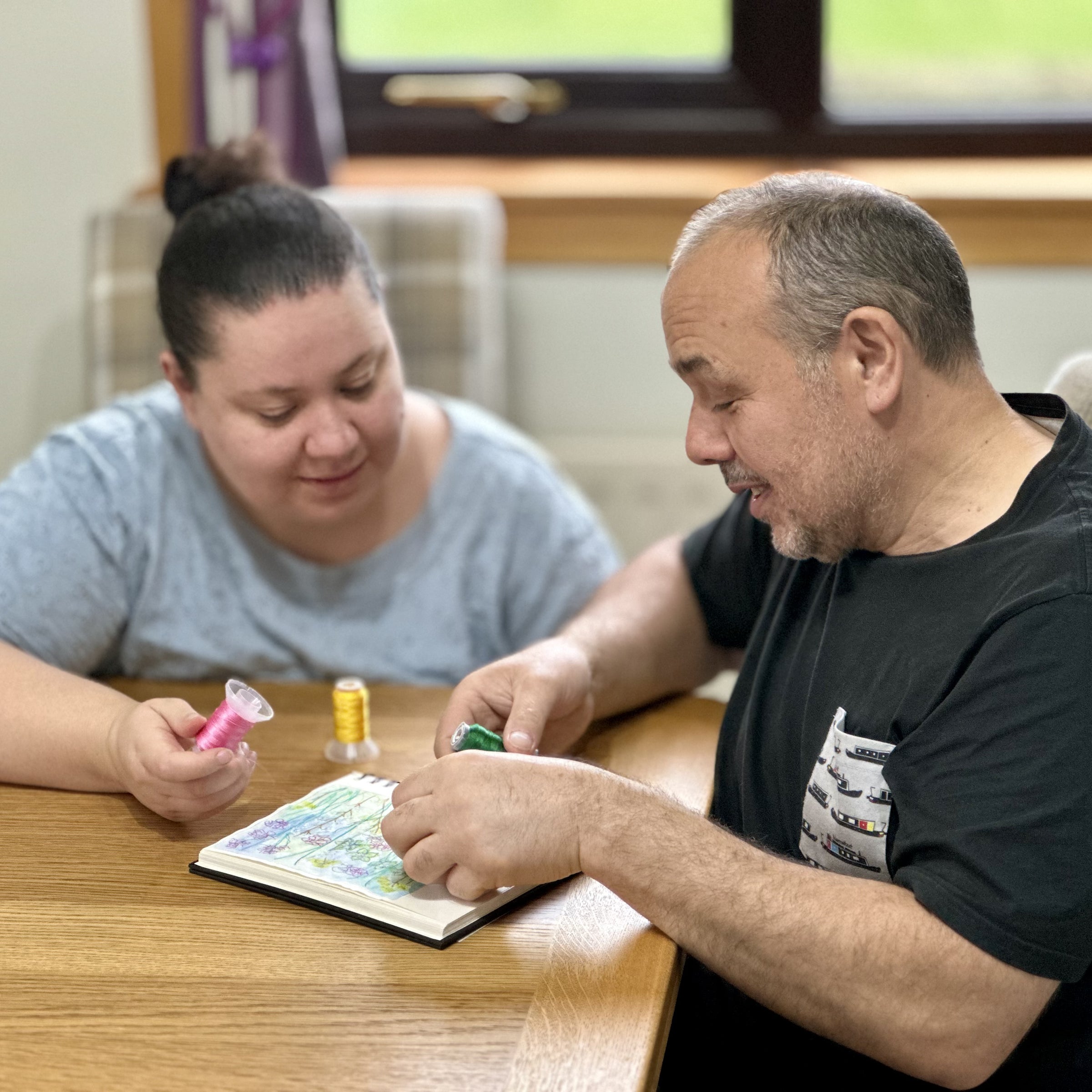Two people sitting at a table with a book and markers, possibly engaged in an educational activity.