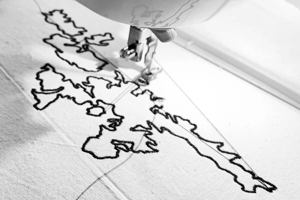 A close-up black and white photo of a sewing machine needle moving over a white fabric surface, creating a stitched outline pattern.