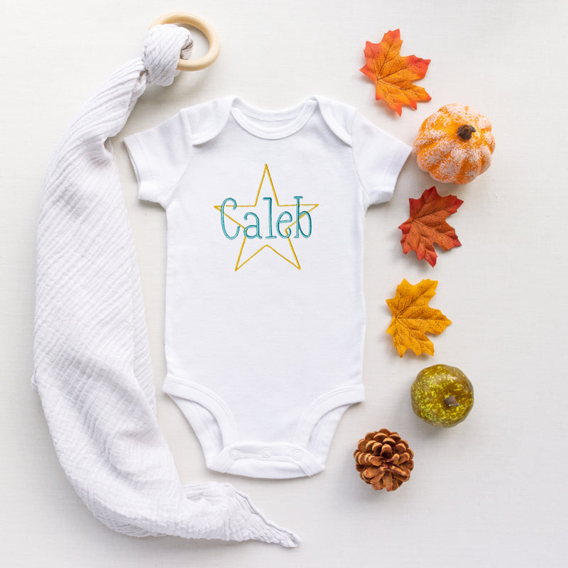 White baby onesie with 'Caleb' printed on a star, surrounded by colorful leaves and pumpkins on a light background