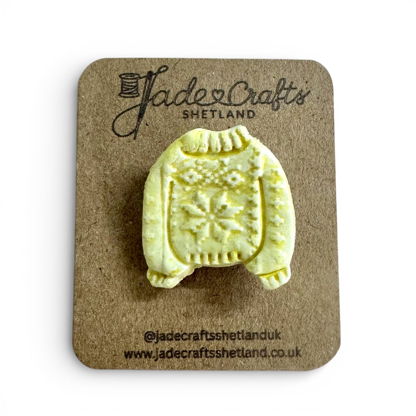 Hand-painted yellow clay brooch in the shape of a Fair Isle jumper on a brown cardboard backing.