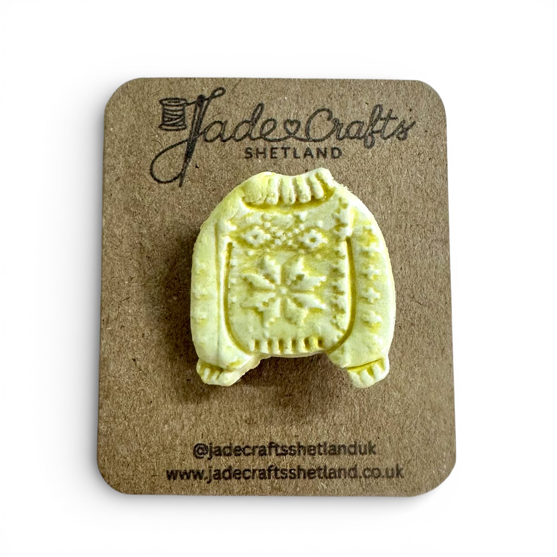 Hand-painted yellow clay brooch in the shape of a Fair Isle jumper on a brown cardboard backing.