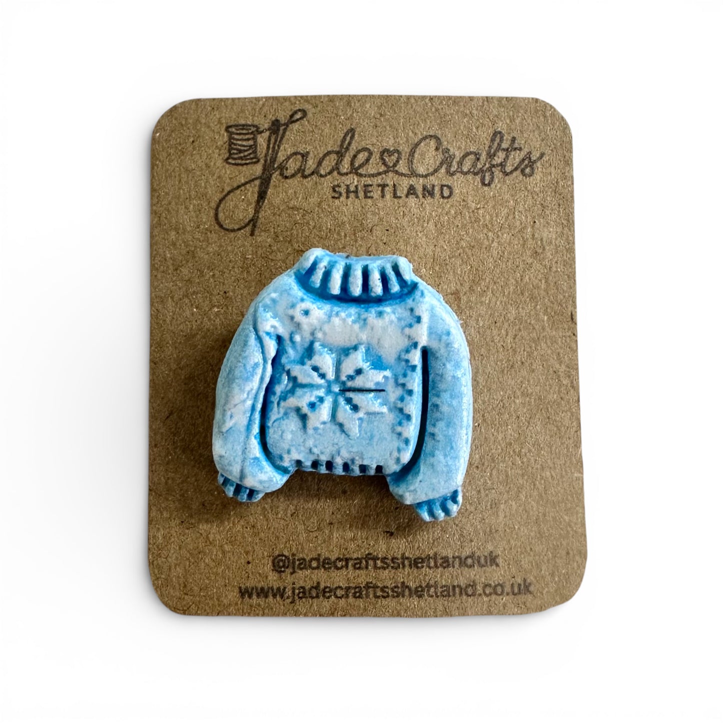 Hand-painted blue clay brooch in the shape of a Fair Isle jumper on a brown card backing.