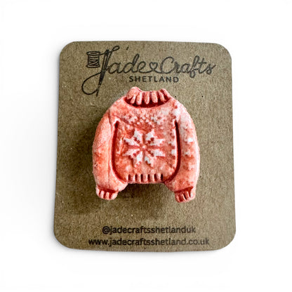 Hand-painted clay brooch in the shape of a red Fair Isle jumper on a brown cardboard backing.